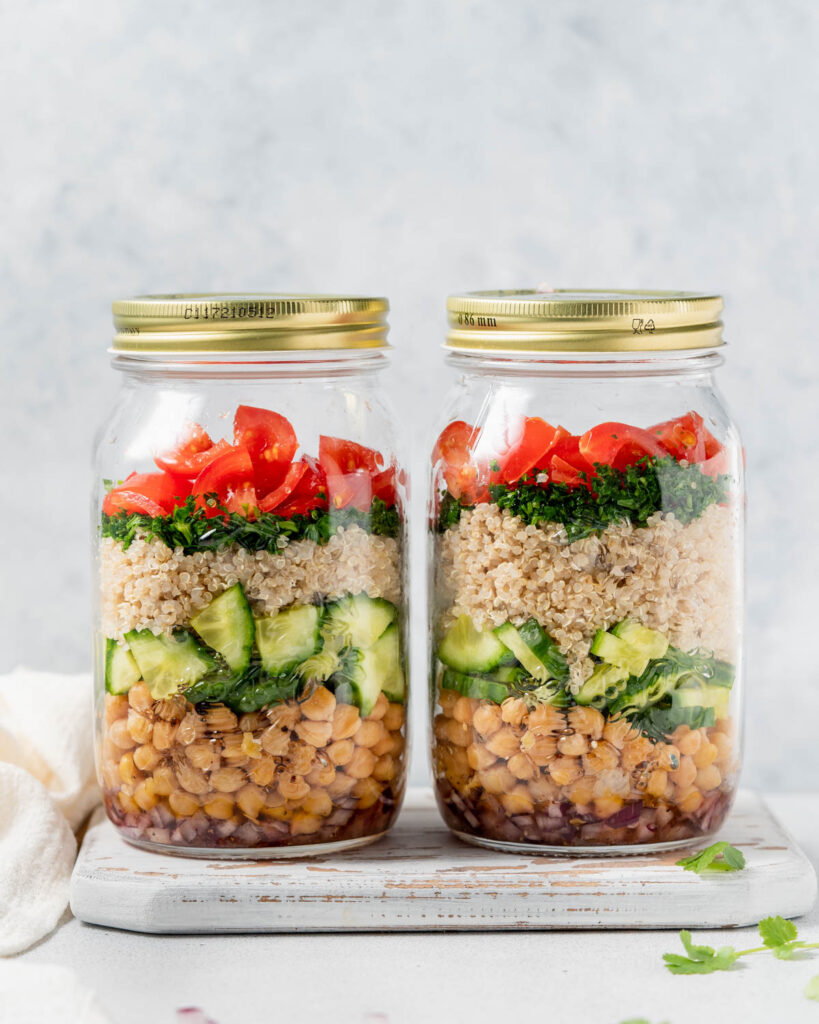 Two salad jars with chickpeas, cucumber, quinoa, herbs and tomatoes layered in mason jars.
