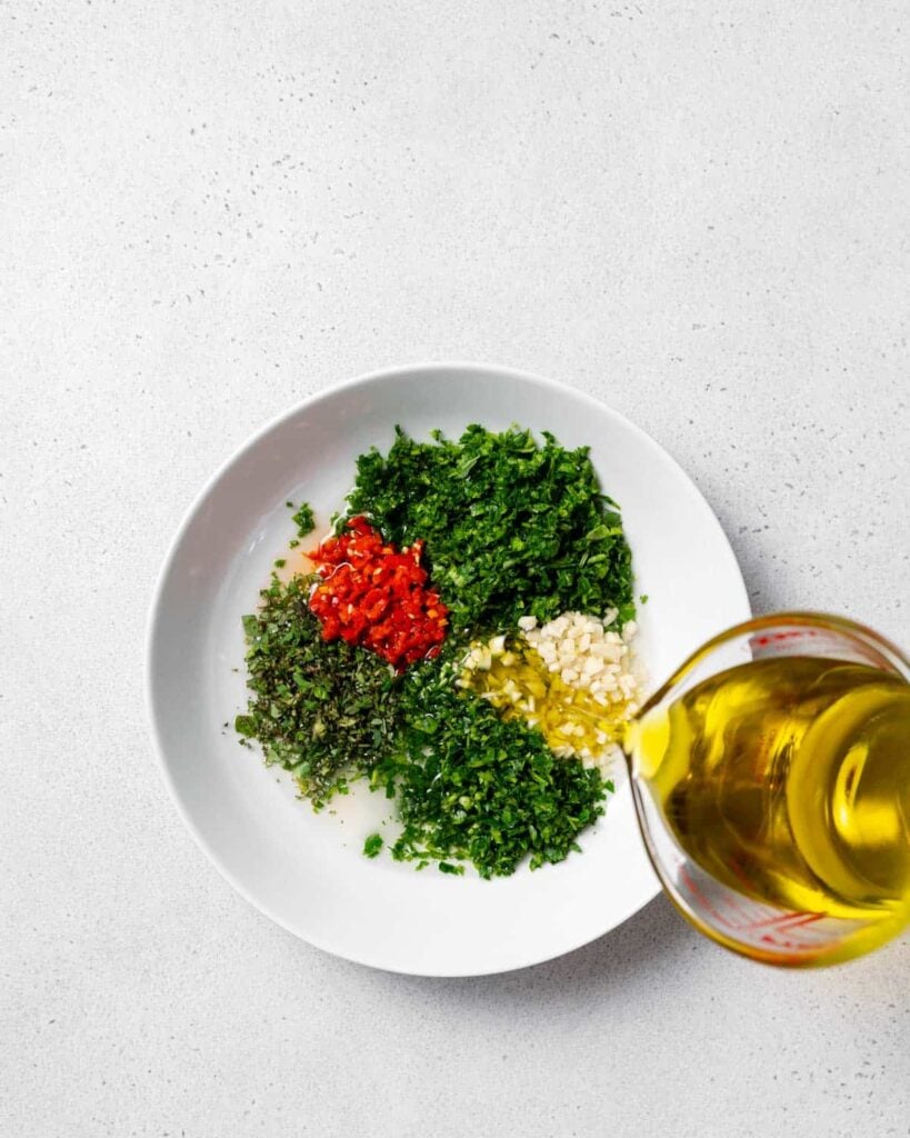 olive oil being poured into a bowl with cilantro peppers and ingredients in a bowl to make chimichurri sauce 