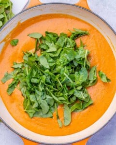 Chopped fresh baby spinach sitting on top of smooth red pepper sauce in a skillet, ready to stir in.