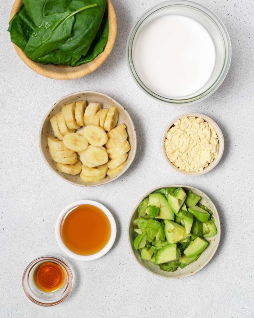 Spinach leaves, sliced banana, avocado chunks, milk, honey and protein powder divided into small bowls.