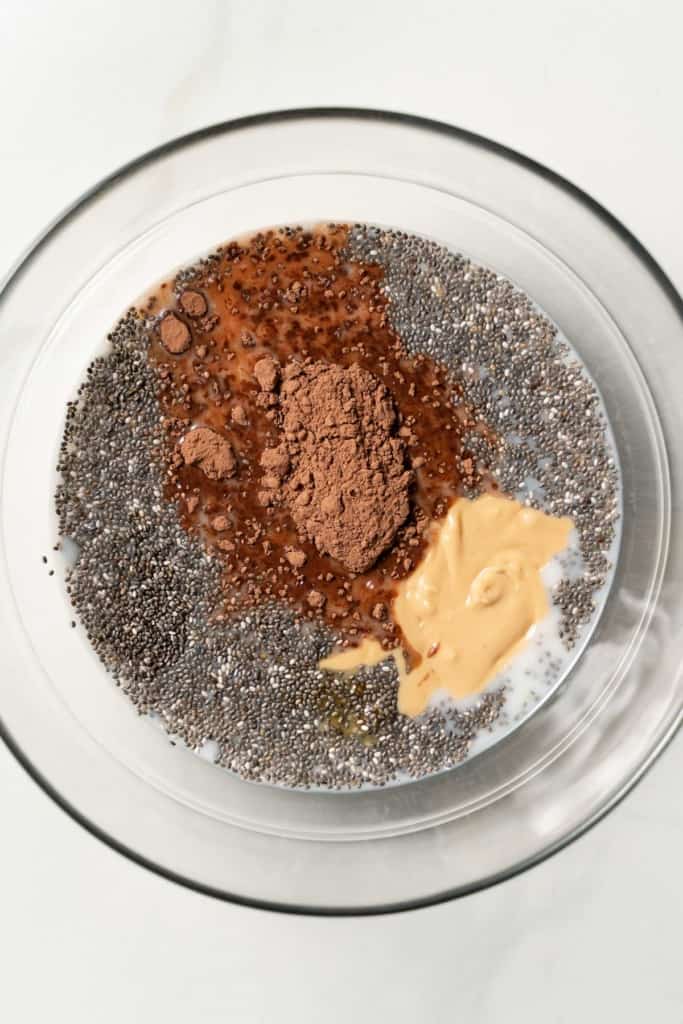 Milk, chia seeds, peanut butter and cacao powder added to a glass mixing bowl.