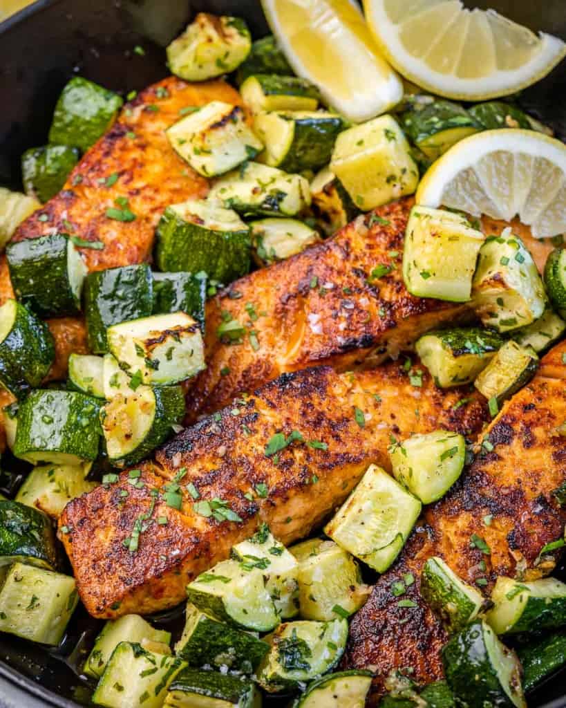 close up image of baked salmon filet on a black skillet with chunks of zucchini.
