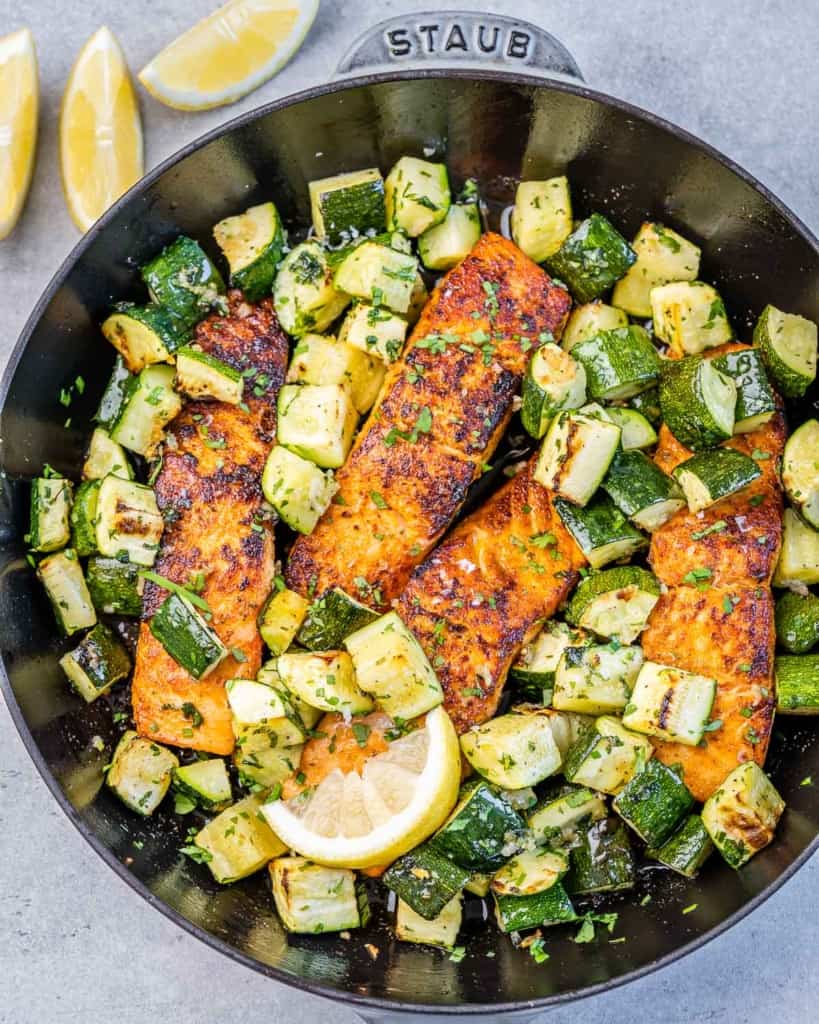 top view salmon filets cook on a black skillet with chunks of zucchini