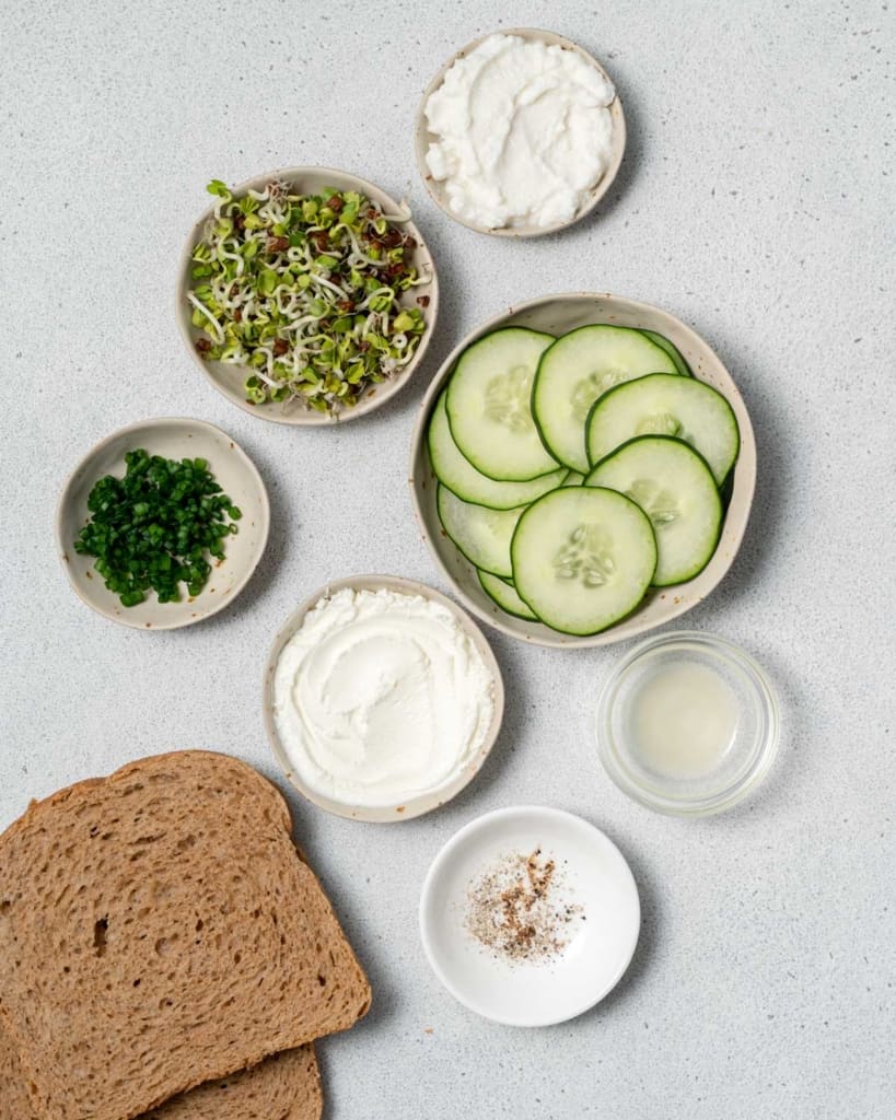 Cream cheese, ricotta cheese, sliced cucumbers, bread and sprouts divided into small portions.