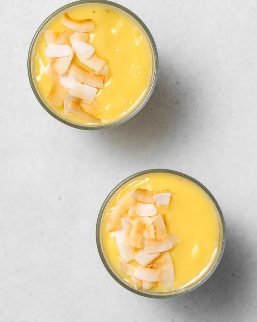 Two mango smoothies in glasses, topped with coconut flakes.