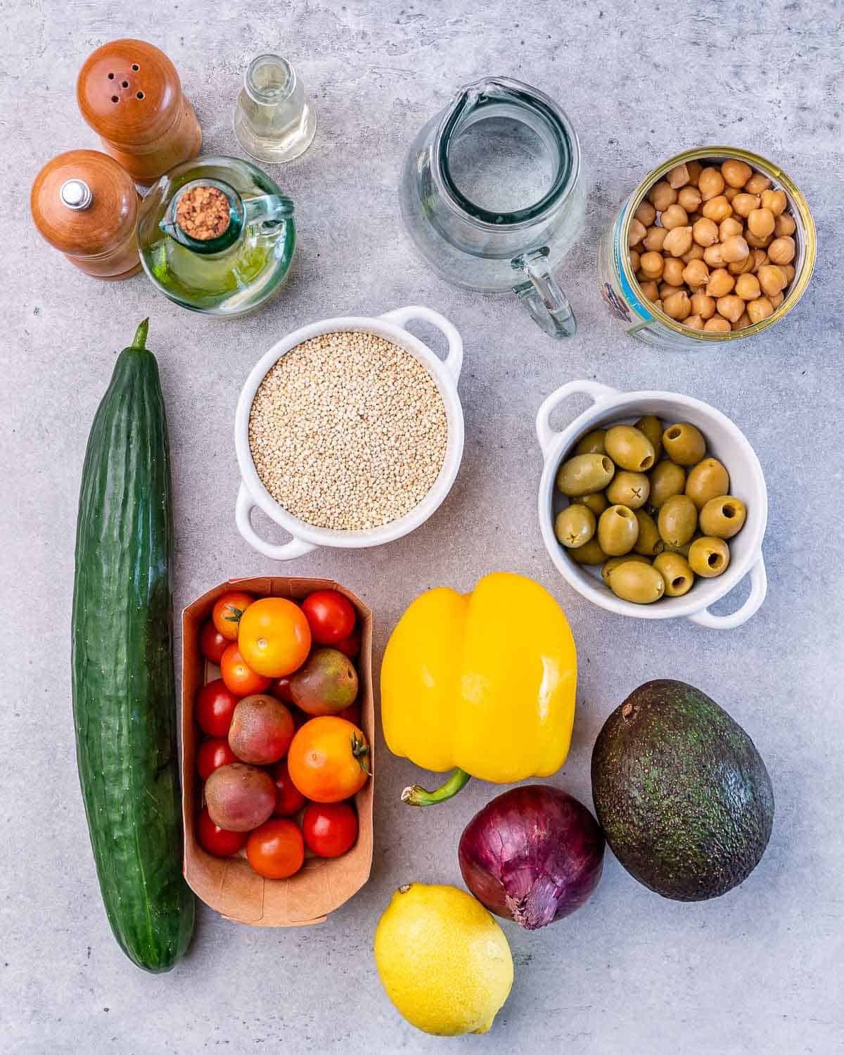 A bowl of uncooked quinoa near a cucumber, cherry tomatoes, an avocado, onion, bell pepper, a lemon and a bowl of green olives.