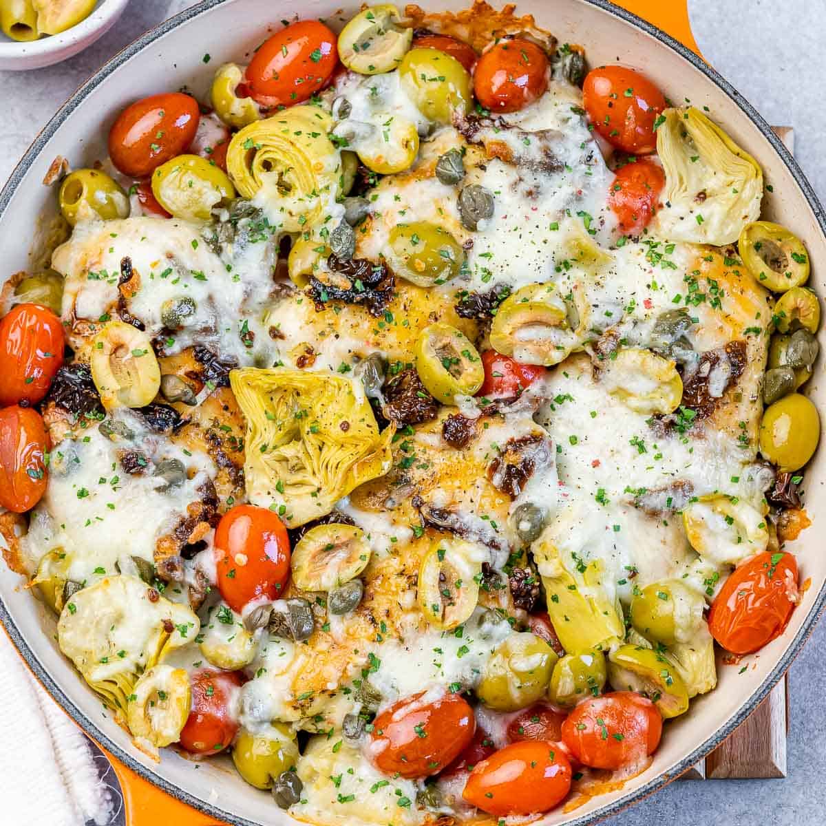 top view of baked chicken breast in a skillet with cherry tomatoes, olives, artichokes and melted cheese