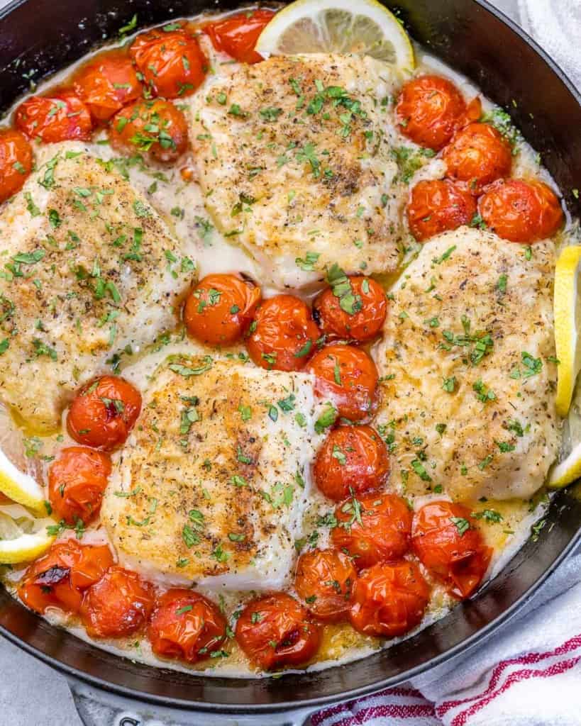 close up image of 4 baded cod fillets on a black pan with lemon garnished and cooked cherry tomatoes around the cods  