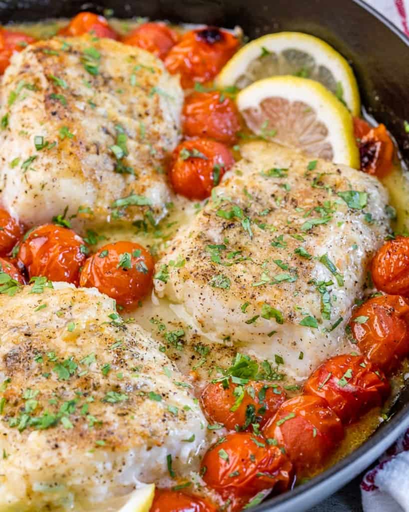 close up image of cooked cod with blistered tomatoes and lemon wedges to garnish