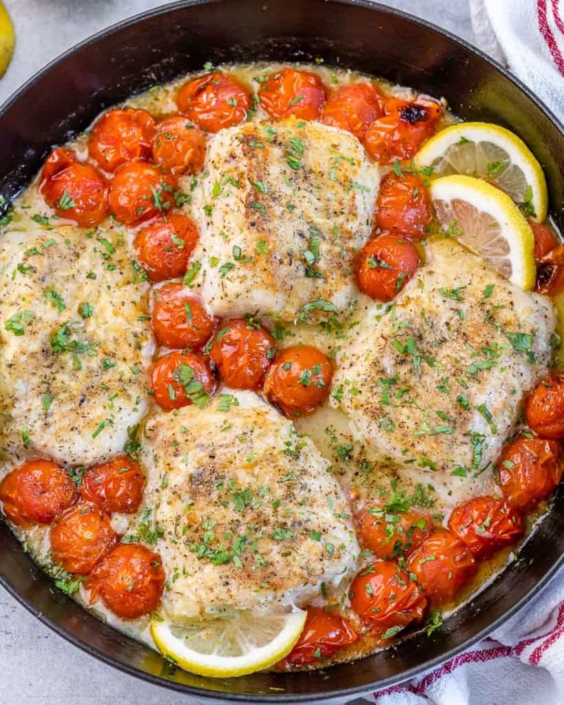top view of a black skillet with 4 baked cod fillets and blistered cherry tomatoes along with lemon slices garnishes