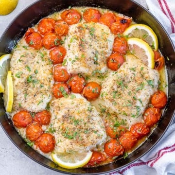 top view of a round black skillet with 4 baked cods, blistered tomatoes and lemon slices