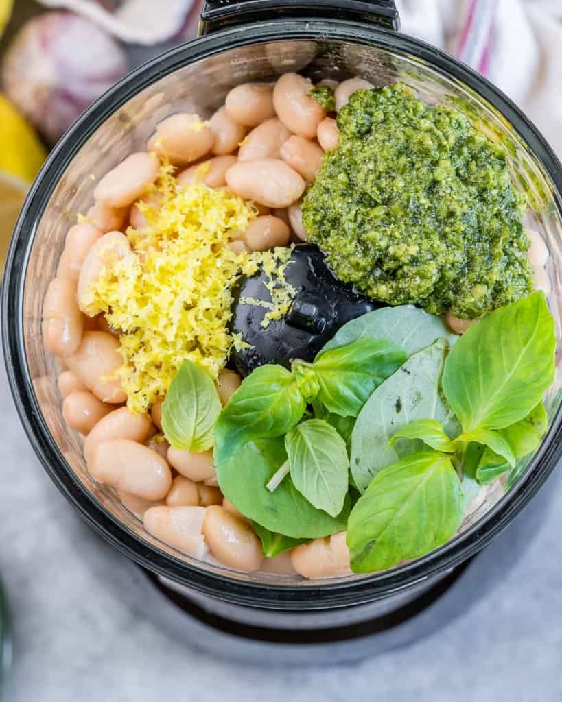 Adding white beans, pesto and basil to a food processor.