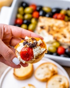 hand holding a baguette topped with feta cheese dip and baked tomatoes and olives