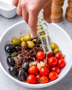 hand holding a jar of oil being poured over tomatoes and olives in a white bowl