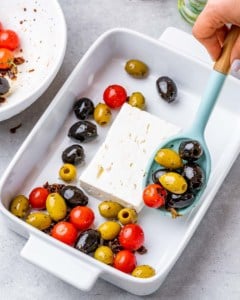 feta cheese block in a white dish with hand holding spoon and adding olives and tomato mixture
