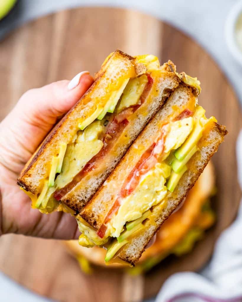 Hand holding a sandwich made with eggs, cheese, avocado and tomato.