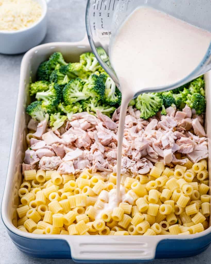 Pouring Alfredo sauce over chicken, pasta and broccoli.