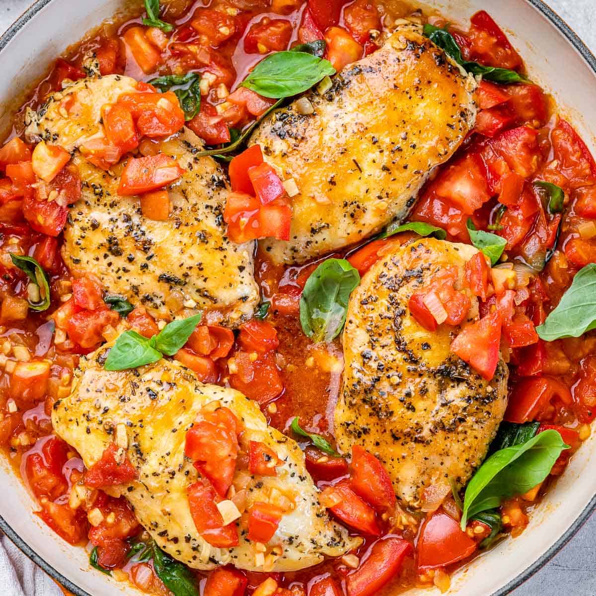 top view of 4 cooked seasoned chicken breasts in a red tomato base sauce and basil garnishes on an orange skillet