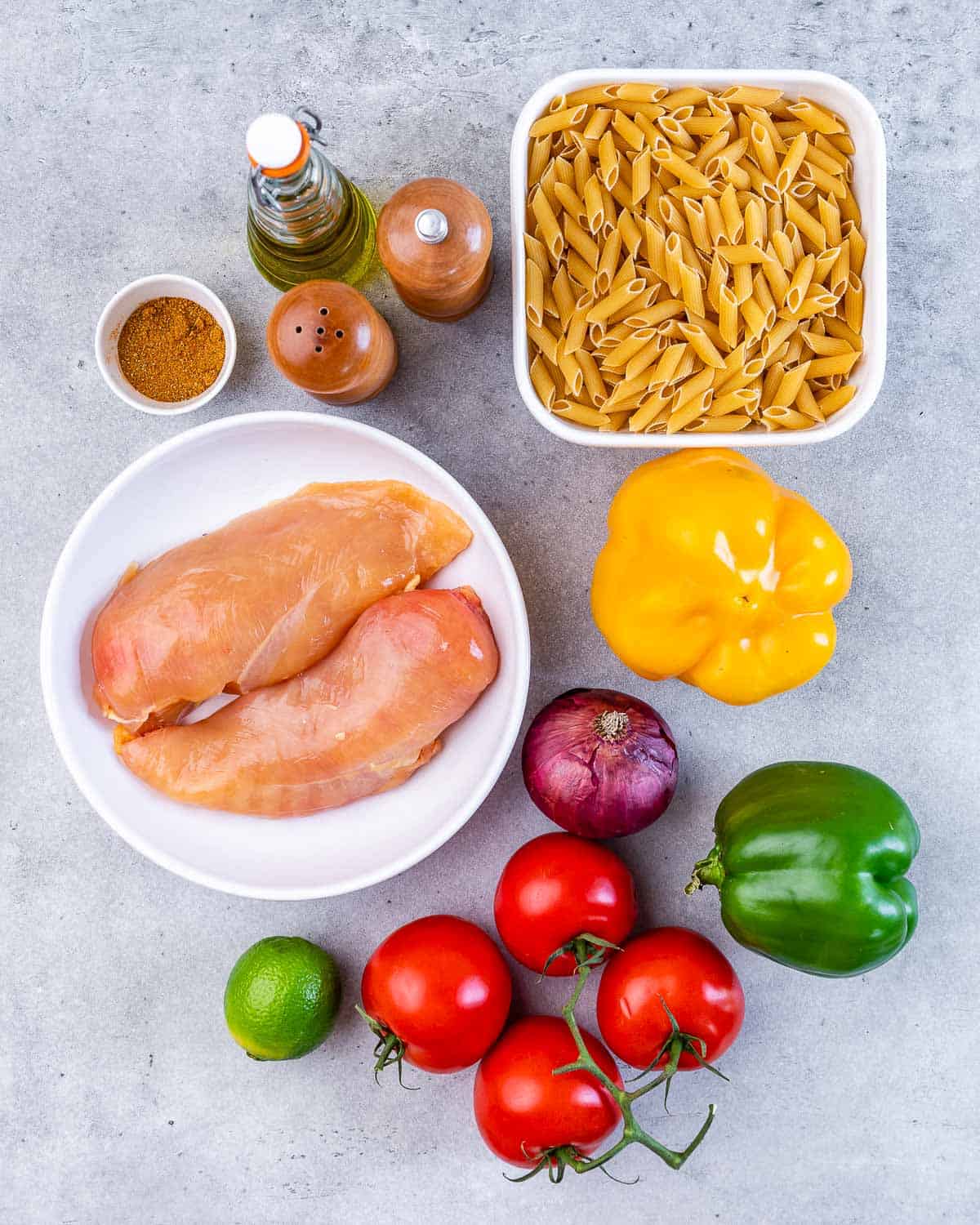 Raw chicken breasts on a plate near a bowl of pasta and tomatoes, a lime and bell peppers.