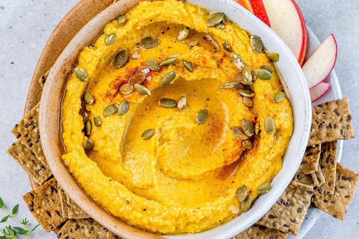 Top view of round white bowl with pumpkin hummus thats orange in color topped with pumpkin seeds and a side of crackers by the bowl.