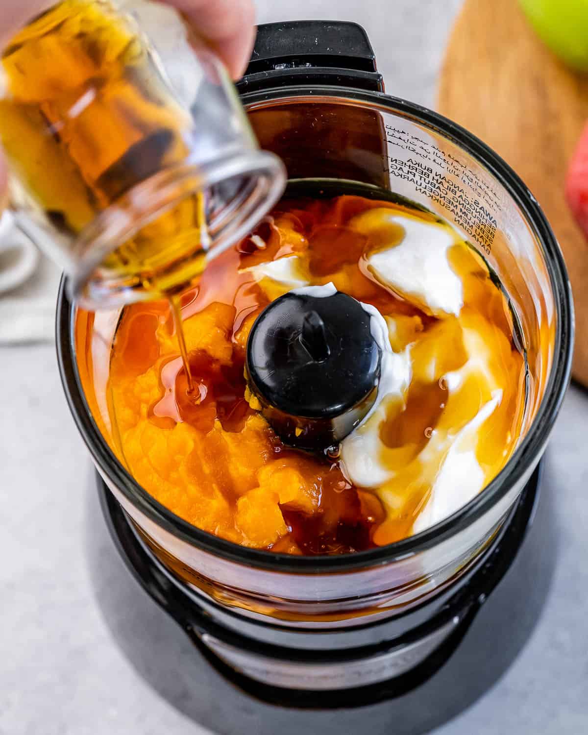 Pouring maple syrup into a food processor with pumpkin and greek yogurt.
