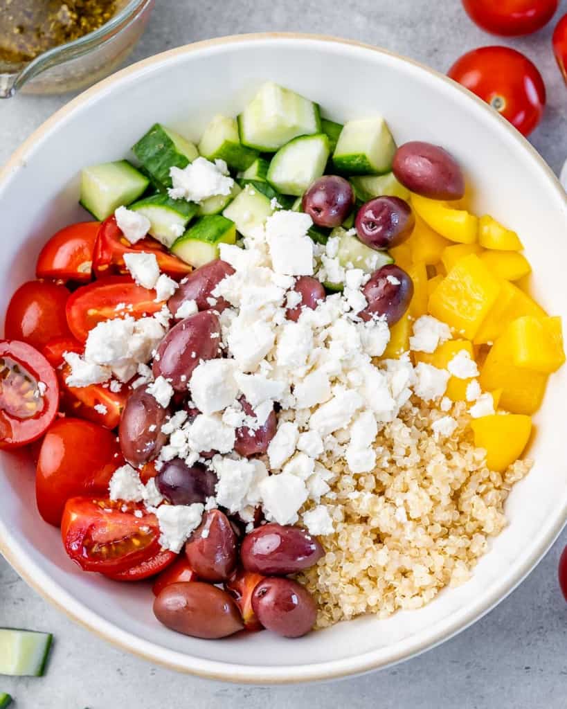chopped veggies, quinoa, olives, and crumbled feta cheese added in a white bowl