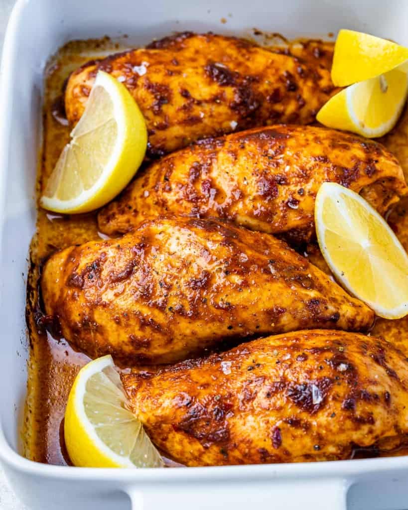 close up of 4 baked chicken breasts that are orange looking in a white baking dish with lemon garnishes
