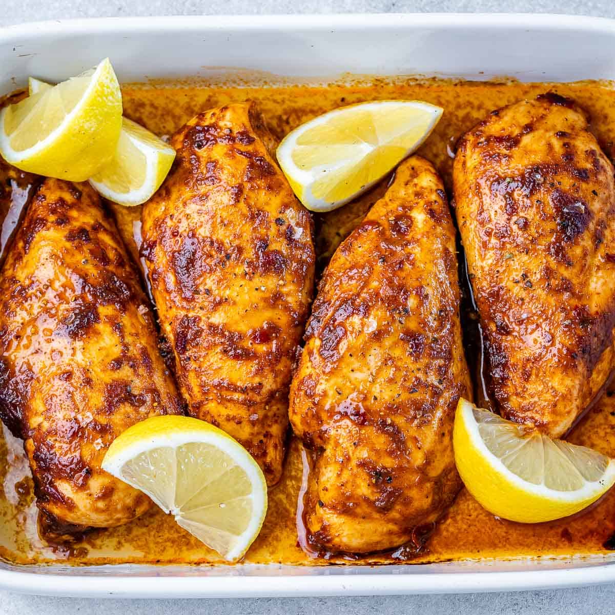 top view of 4 baked chicken breasts that are orange in color in a white dish with lemon wedges as garnishes