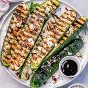 grilled zucchini halves on a round white plate drizzled with balsamic vinegar and goat cheese.