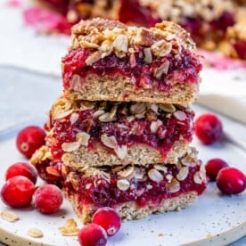 3 oatmeal bar squares filled with cranberry jam and they are stacked over each other on a plate with whole cranberries by the bars for styling.