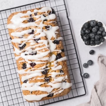 top view of a baked blueberry bread with icing drizzled on top on a wire wrack and a side of fresh blueberries in a small bowl