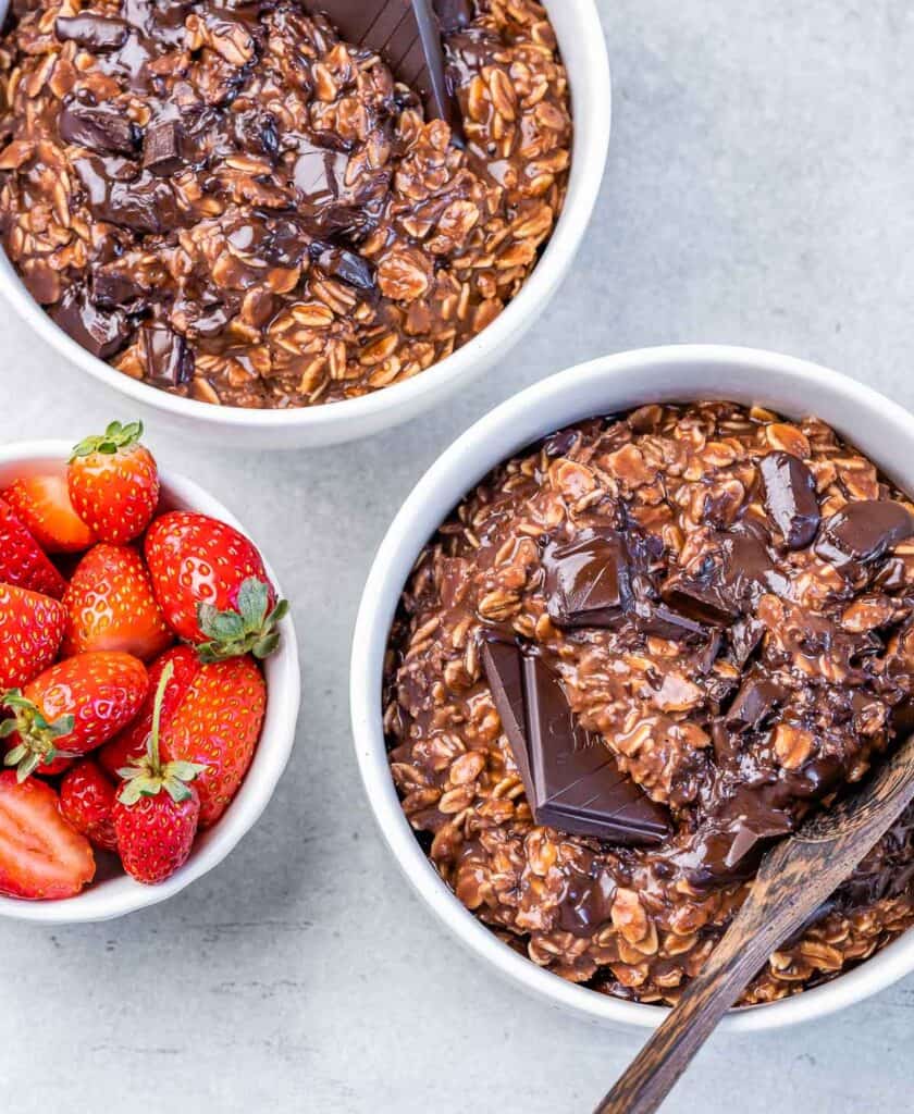 top view of 2 bowls of chocolate oatmeal  with wooden spoon in one of the bowls and a bowl of fresh strawberries 
