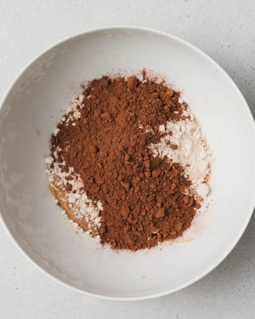 Adding cocoa powder and almond flour to a white bowl.