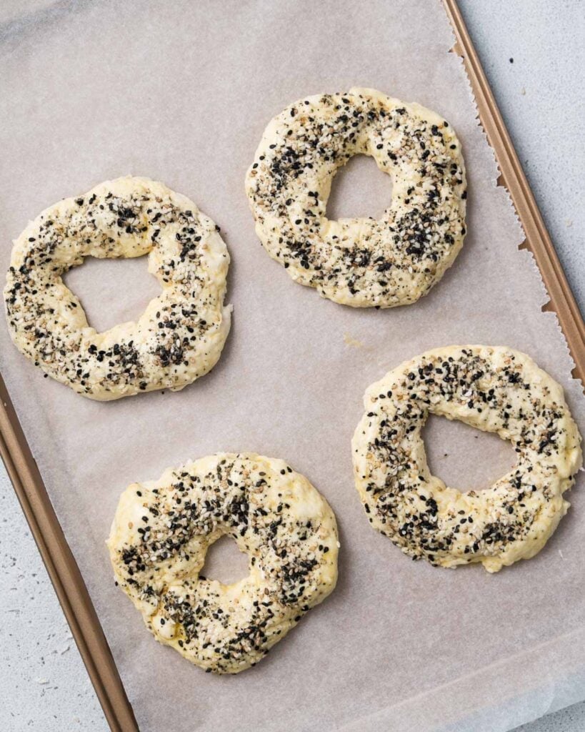 4 unbaked bagel dough added on a sheet pan lined up with parchment paper 