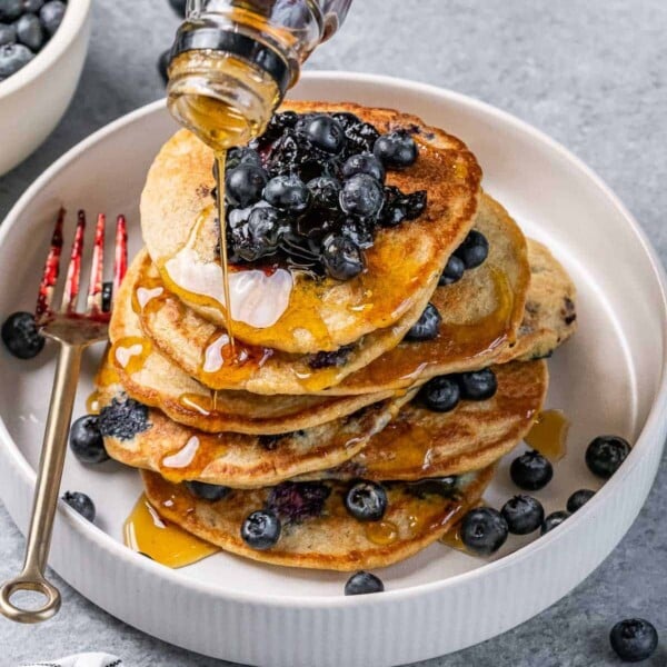 Pouring syrup over a stack of pancakes with blueberries.