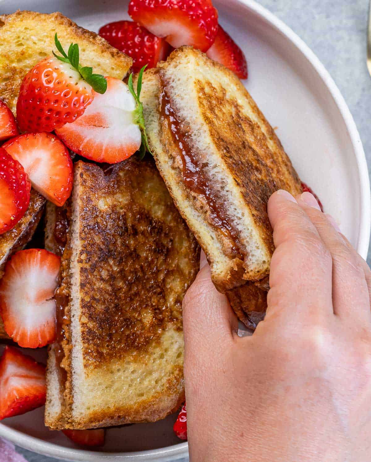 Hand picking up a peanut butter jelly French toast near sliced strawberries.