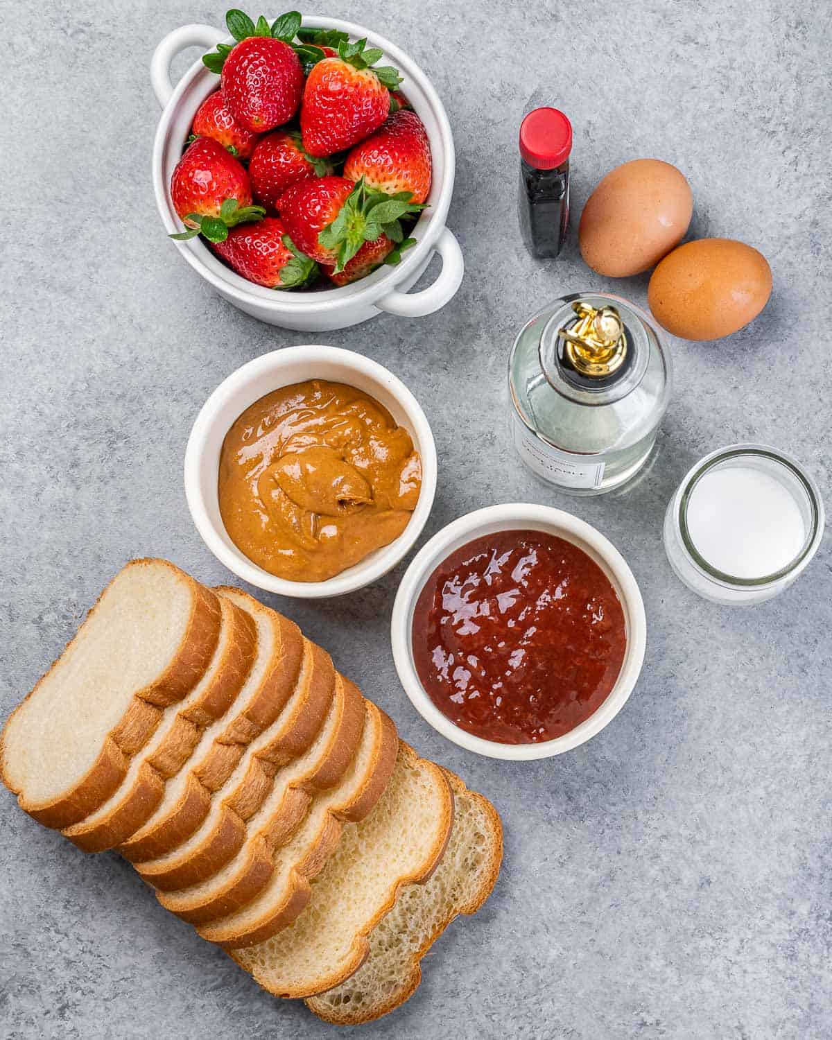Slices of bread, bowls of peanut butter, jelly and strawberries near two eggs, milk and vanilla.