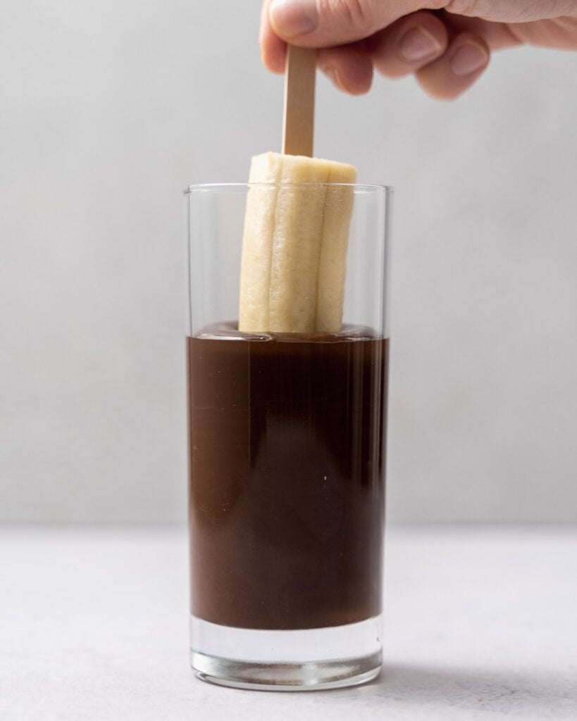 banana pop being dipped into a cup of melted chocolate