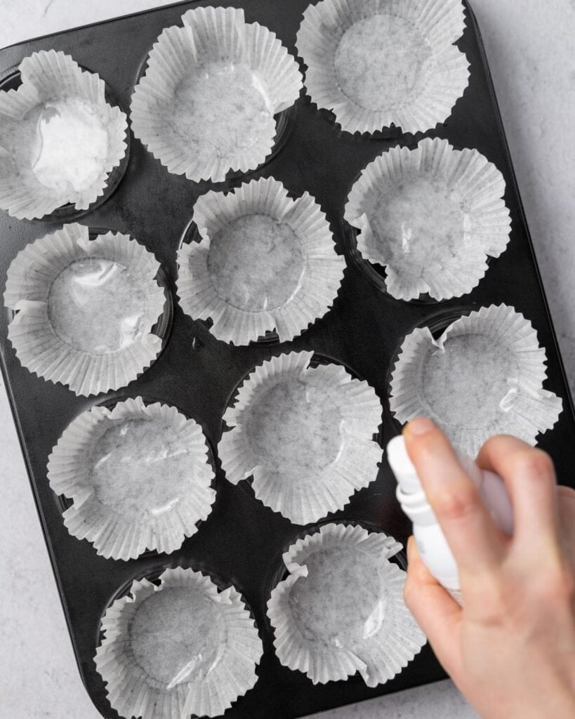 Adding cupcake liners to a pan and spraying them with cooking spray.