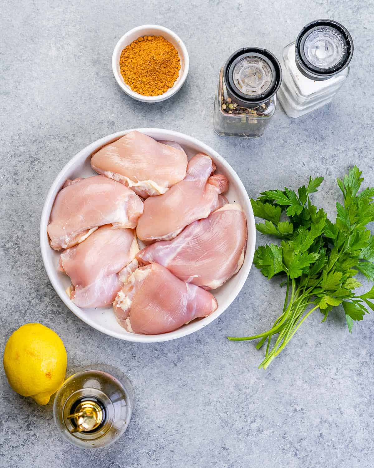 Raw chicken thighs in a bowl near fresh parsley, a lemon, olive oil and a small bowl of taco seasoning.