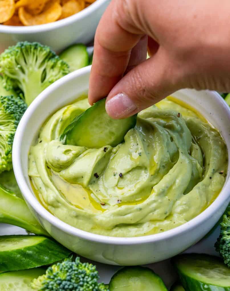 hand holding a peace of cucumber and dipping in the avocado rand dip in a bowl 