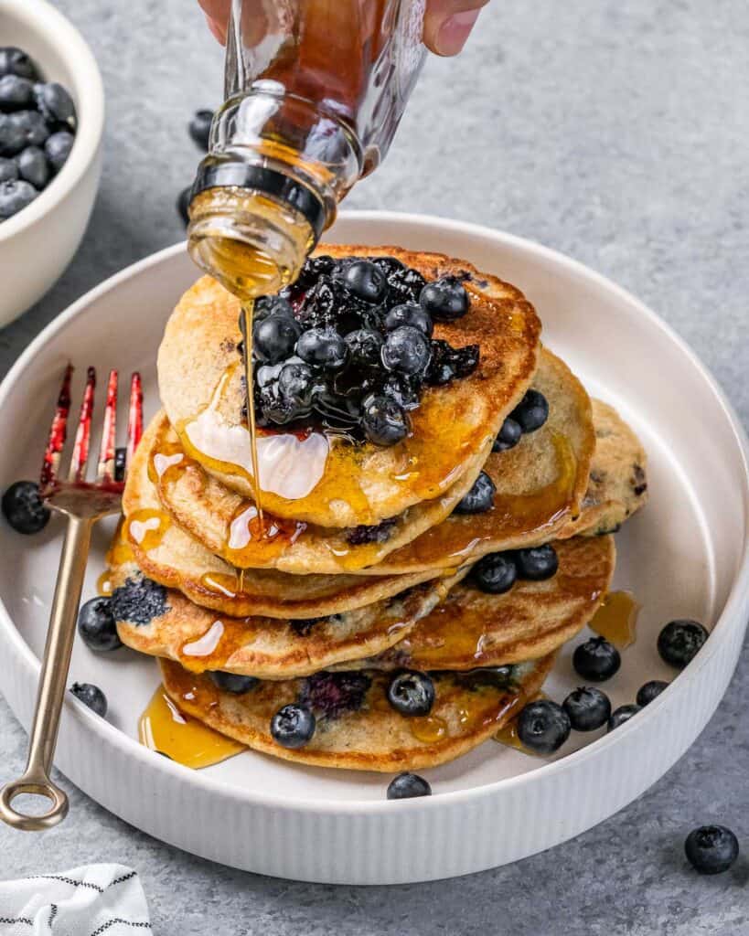 maple syrup being added over blueberry pancakes on a plate