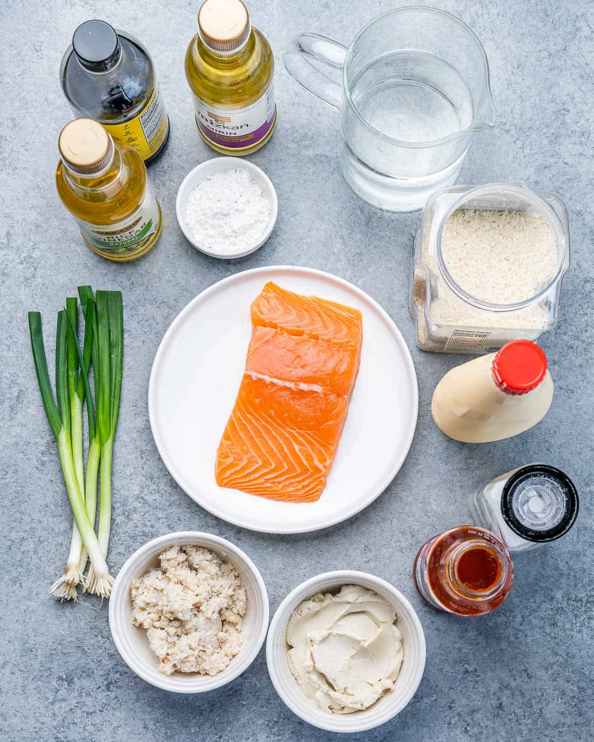 Raw salmon on a plate near green onions, bowls of mayo and cream cheese, and seasoning like rice vinegar, soy sauce and sriracha sauce.