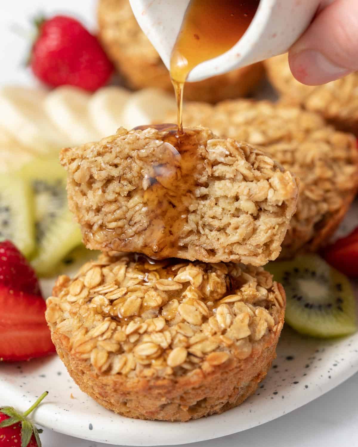 Maple syrup being added over half of a muffin stacked over another whole muffin on a plate