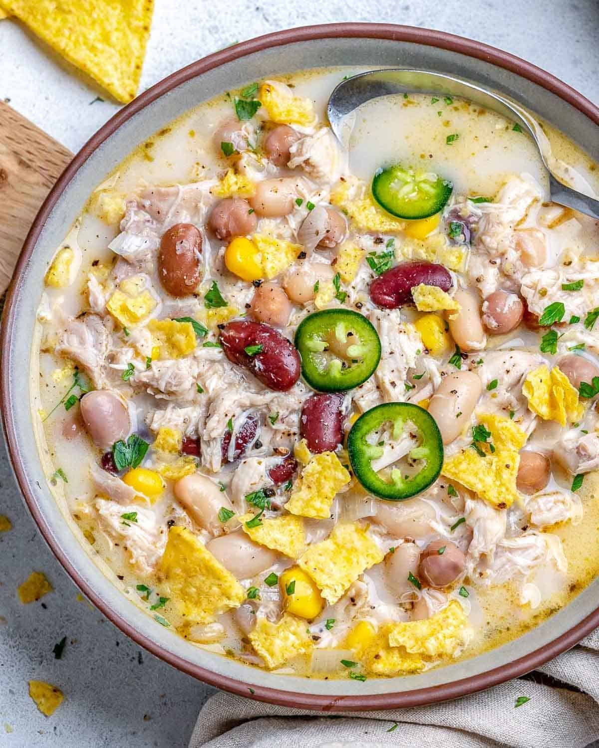 a bowl with white chicken chili made with 4 beans, topped with sliced jalapenos, and crushed tortialla.