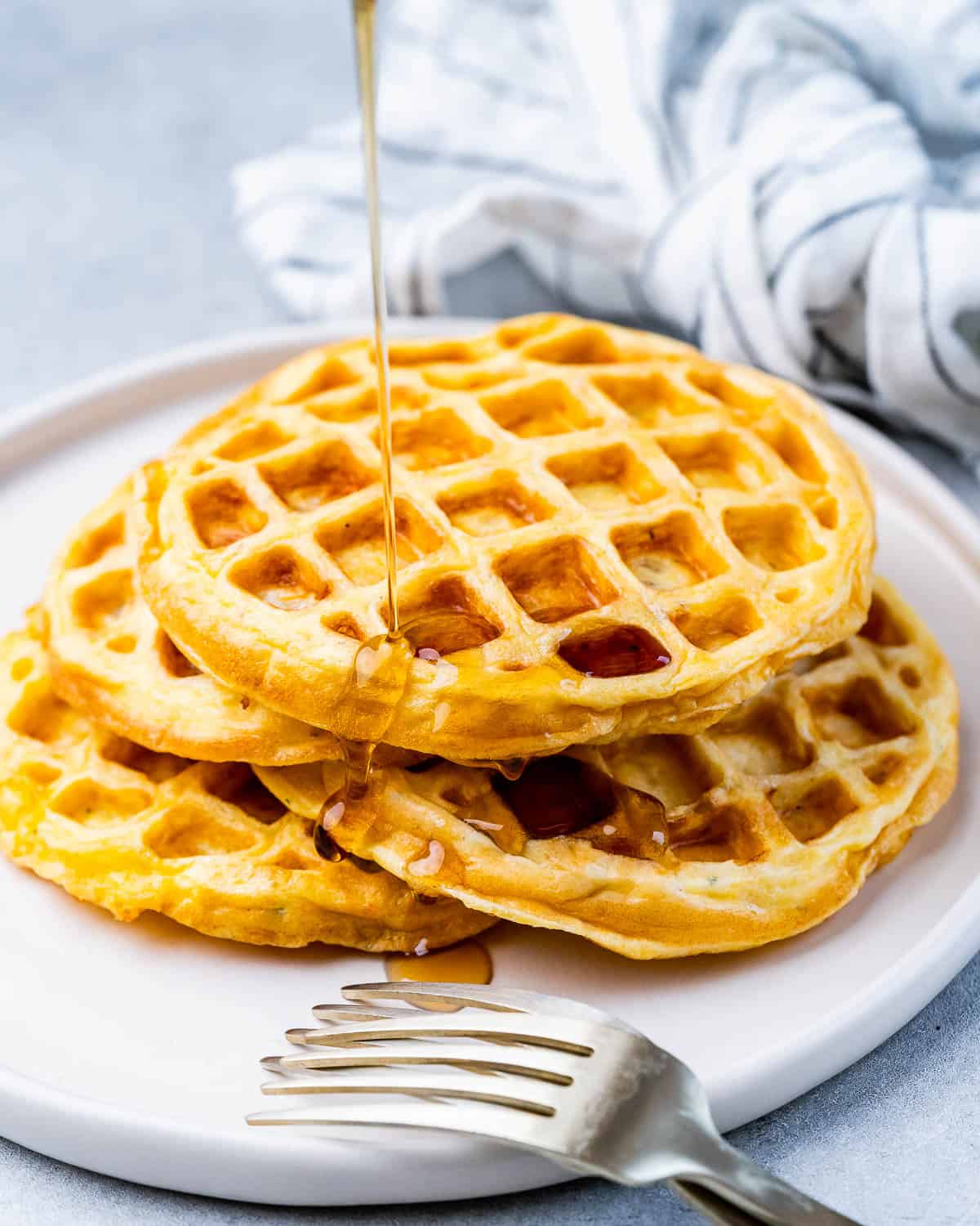 Three warm chaffles stacked on a white plate, with syrup being poured over the top.