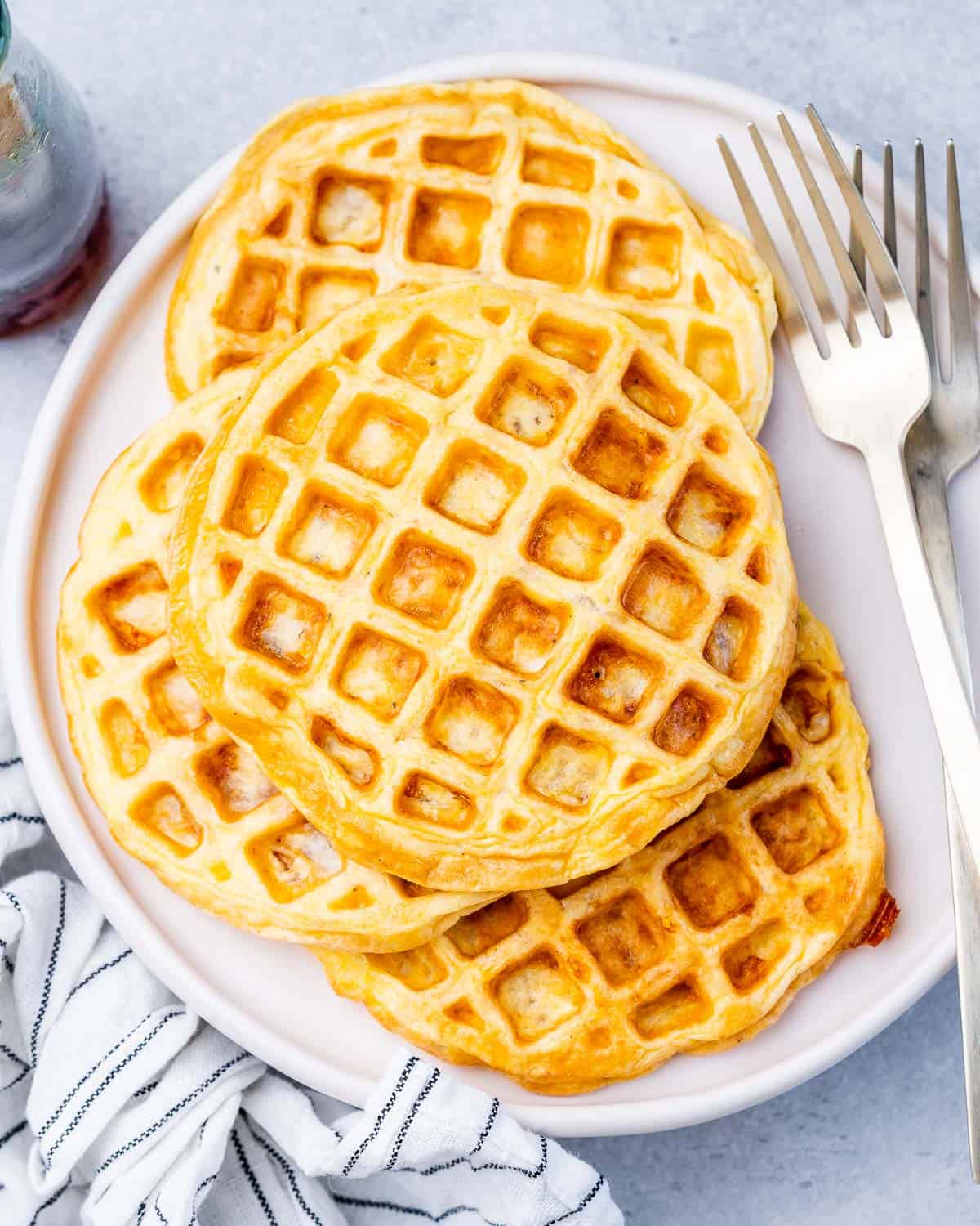 A full plate of freshly cooked chaffles next to two silver forks and a striped cloth napkin.