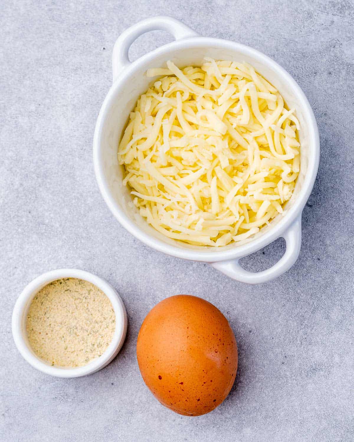 Overhead view of a white bowl filled with shredded mozzarella, a small ramekin of garlic salt, and one whole egg.