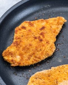 Breaded chicken cutlet cooking in a skillet until golden brown and crispy on the edges.