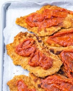 Close-up of crispy golden chicken cutlets topped with dollops of marinara sauce on a baking tray.